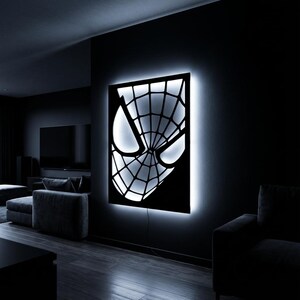 May include: A rectangular wall-mounted light fixture featuring a black cutout of a superhero mask with a web pattern. The design is illuminated from behind by a bright white LED glow, creating a high-contrast silhouette against the dark wall.