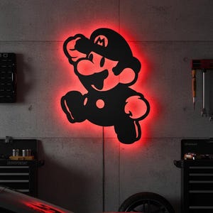 K&ouml;nnte beinhalten: Schwarze Silhouette der Videospiel-Figur Mario, mit roter Neonbeleuchtung. Das Kunstwerk ist an einer Betonwand befestigt und erzeugt einen auff&auml;lligen Kontrast. Das Bild zeigt einen springenden Mario.