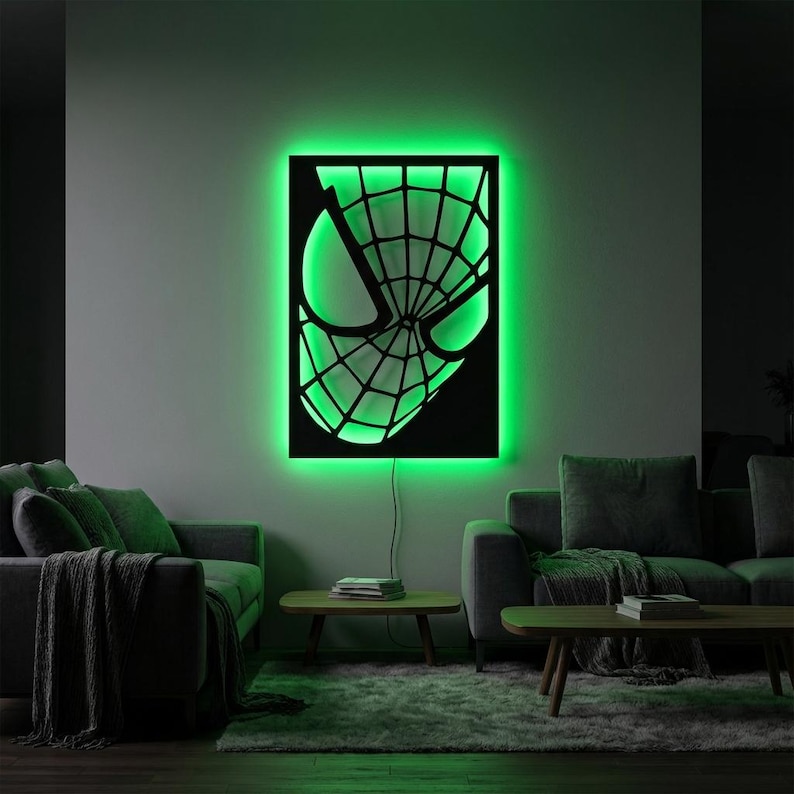 May include: A rectangular black wall art piece featuring a cutout of a superhero mask with a web pattern, illuminated from behind by a vibrant green LED light that casts a glow against the wall, with a thin power cord extending downward.