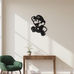 K&ouml;nnte beinhalten: Schwarze Metall-Wandkunst eines rennenden Zeichentrickcharakters mit einem Hut mit einem "M" darauf. Das Kunstwerk ist an einer wei&szlig;en Wand angebracht. Ein gr&uuml;ner Stuhl, ein Holztisch und eine Vase mit wei&szlig;en Blumen befinden sich im Raum.