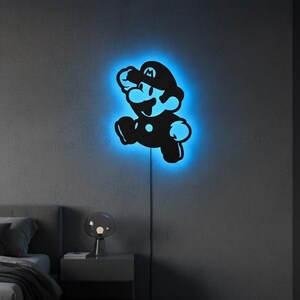 K&ouml;nnte beinhalten: Schwarze Silhouette der Videospiel-Figur Mario in Laufpose, mit blauem Neonlicht, das die Figur umrandet. Die Wandkunst ist an einer grauen Wand angebracht, mit einem Netzkabel sichtbar.