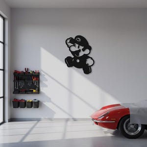 K&ouml;nnte beinhalten: Schwarze Metall-Wandkunst des Videospielcharakters Mario in einer Laufpose. Das Kunstwerk ist an einer wei&szlig;en Wand in einer Garage angebracht. Ein roter Sportwagen ist teilweise im Hintergrund zu sehen.