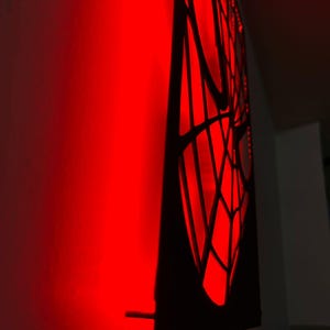 May include: A black wall-mounted panel featuring a cutout spiderweb design illuminated from behind by a bright red light. The light casts a vibrant red glow onto the adjacent wall surface, highlighting the intricate web pattern in silhouette.