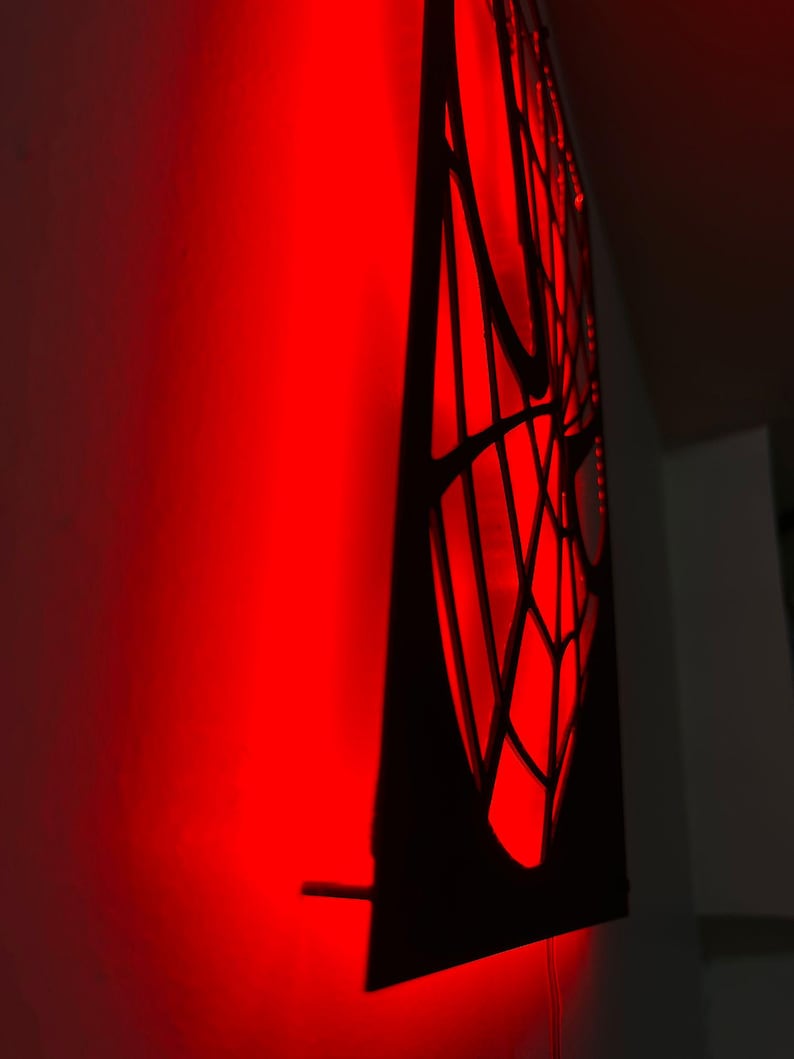 May include: A black wall-mounted panel featuring a cutout spiderweb design illuminated from behind by a bright red light. The light casts a vibrant red glow onto the adjacent wall surface, highlighting the intricate web pattern in silhouette.