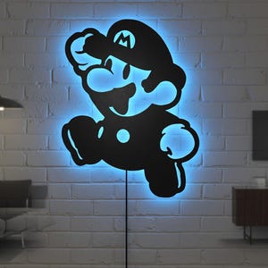 K&ouml;nnte beinhalten: Schwarze Silhouette der Videospiel-Figur Mario, beleuchtet von einem blauen LED-Licht. Das Licht ist an einer wei&szlig;en Backsteinmauer befestigt, mit einem Netzkabel, das nach unten verl&auml;uft.