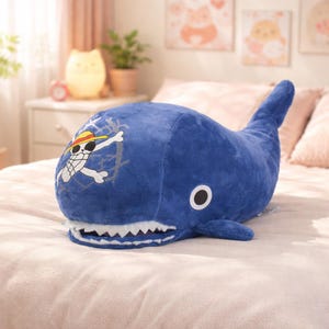 May include: A plush, blue whale-shaped pillow with a white and yellow pirate logo on its side. The pillow has a white, toothy grin and a single black eye. The whale's tail is upright. The pillow is on a bed.