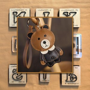 May include: A brown leather teddy bear keychain with a white face and black accents. The bear is wearing a black jacket with a white collar. The keychain is attached to a brown leather strap. The background includes wooden blocks with text.