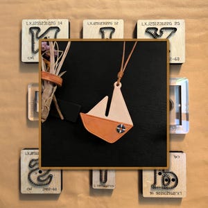 Könnte beinhalten: Lederanhänger in Segelbootform mit beigem Segel und orangefarbenem Rumpf, verziert mit einem blau-weißen Emblem. Der Anhänger hängt an einer braunen Kordel. Das Bild ist von Holzblöcken mit graviertem Text umgeben.
