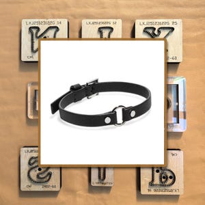 Könnte beinhalten: Schwarze Leder-Halskette mit silberfarbenem O-Ring und Schnallenverschluss. Die Halskette wird auf weißem Hintergrund präsentiert, umgeben von Holzblöcken mit Text und Metallformen. Ca. 30 cm lang.