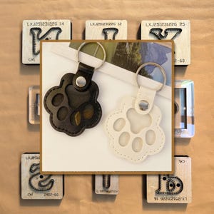 May include: Two paw-shaped keychains, one black and one off-white, with silver key rings. The keychains are on a white background, surrounded by wooden stamps with text and designs. The black keychain is made of leather.