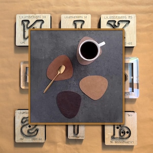 May include: A top-down view showcases a coffee cup on a coaster, a gold spoon, and three leather coasters in shades of brown. The coasters have organic shapes. Surrounding the scene are wooden blocks with metal designs and text.