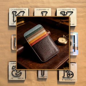 May include: A rectangular leather card holder featuring a dark brown pebbled texture on the main body and a series of horizontal slots in light blue, green, cream, yellow, and orange. The item is positioned against a background of wooden letter dies.
