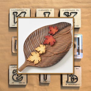 May include: A wooden leaf-shaped serving board with three decorative maple leaves in shades of red and yellow. The board has a textured surface and is surrounded by various wooden stamps with text.