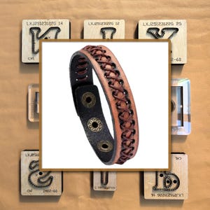 Könnte beinhalten: Ein braunes Lederarmband mit geflochtenem Design und schwarzer Rückseite. Das Armband hat drei messingfarbene Druckknöpfe. Der Hintergrund umfasst verschiedene Holzblöcke mit Text und Metallstempeln.