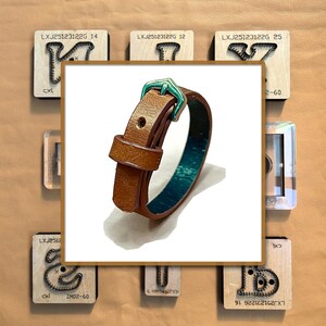 Könnte beinhalten: Ein braunes Lederarmband mit türkisem Innenfutter und einer türkisfarbenen Schnalle. Das Armband wird in einem quadratischen Rahmen präsentiert, umgeben von Holzblöcken mit Text und Metallformen. Ein stilvolles Accessoire.
