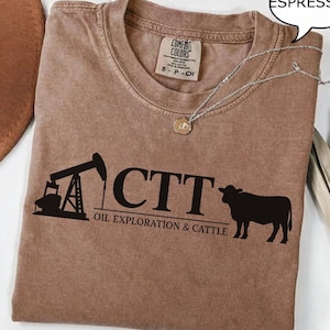 Może przedstawiać: Brązowa koszulka w kolorze espresso z czarną grafiką przedstawiającą wieżę wiertniczą, litery "CTT" i sylwetkę krowy. Pod literami znajduje się napis "OIL EXPLORATION & CATTLE". Na koszulce jest srebrny naszyjnik z wisiorkiem.