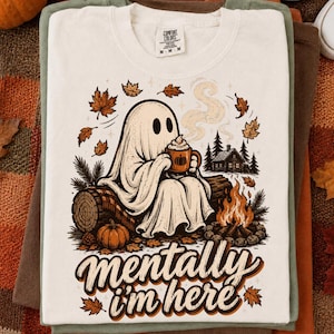 May include: A cream-colored t-shirt featuring a graphic of a ghost sitting on a log, holding a mug with a pumpkin design. The scene includes a campfire, a cabin, and falling autumn leaves. The text "mentally i'm here" is printed in brown script.