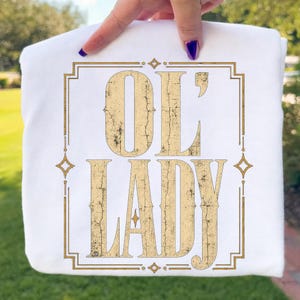 May include: A white t-shirt featuring a gold-colored graphic print with a distressed, cracked texture. The design displays the text "OL' LADY" in a bold, serif font, enclosed within a rectangular border accented with small star-like shapes.