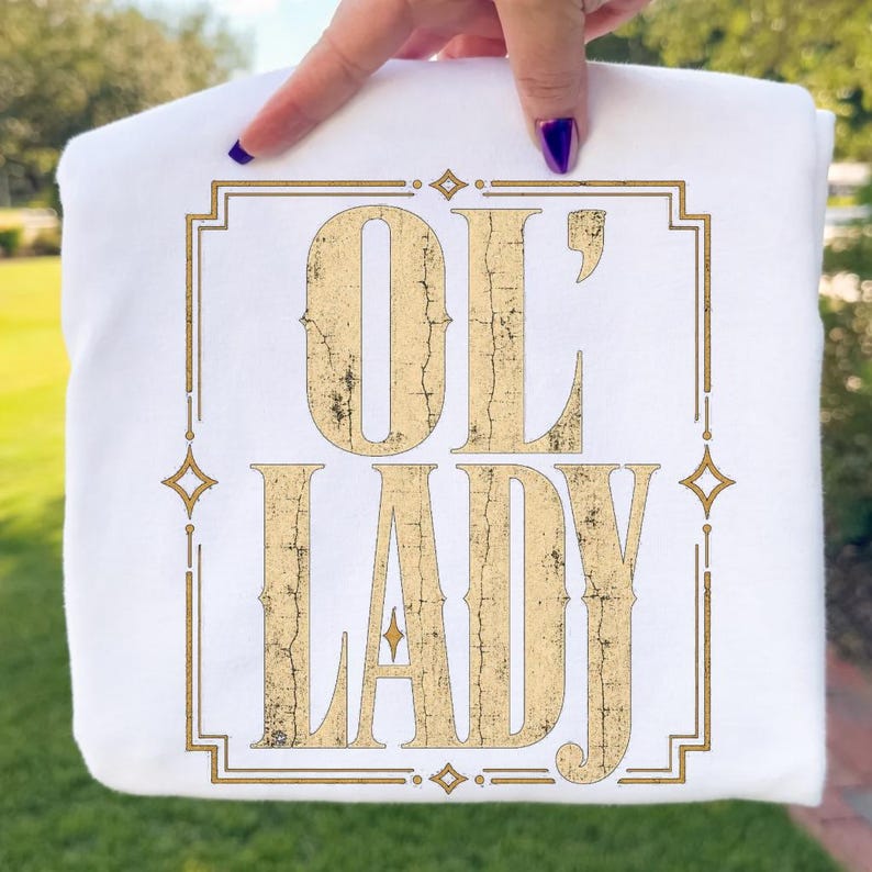 May include: A white t-shirt featuring a gold-colored graphic print with a distressed, cracked texture. The design displays the text "OL' LADY" in a bold, serif font, enclosed within a rectangular border accented with small star-like shapes.