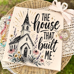 May include: A cream-colored fabric square featuring a black ink illustration of a church with a steeple, surrounded by colorful wildflowers. The text reads, "THE HOUSE THAT BUILT ME" in a bold, black script font with gold star accents.