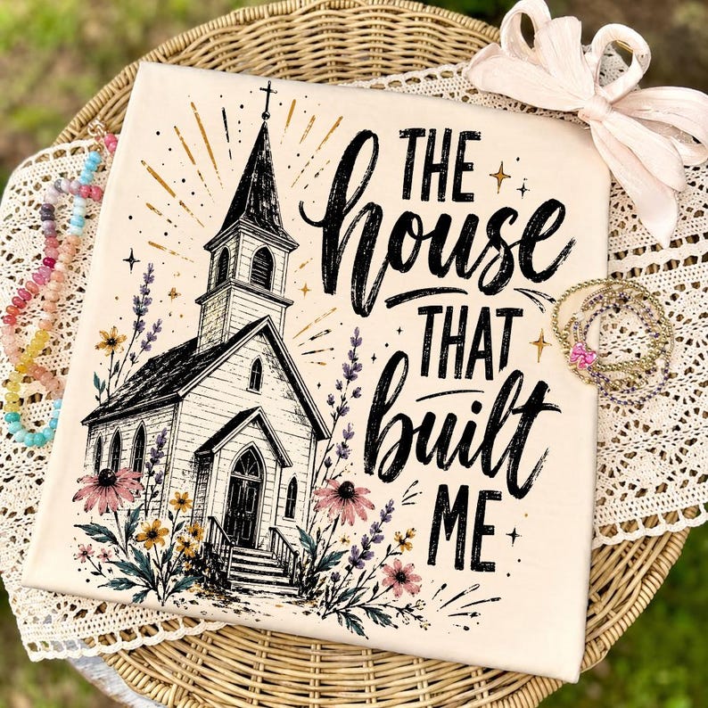 May include: A cream-colored fabric square featuring a black ink illustration of a church with a steeple, surrounded by colorful wildflowers. The text reads, "THE HOUSE THAT BUILT ME" in a bold, black script font with gold star accents.