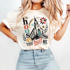 May include: A cream-colored short-sleeve t-shirt featuring a graphic of a church building with the text THE house THAT built ME. The design includes colorful star patterns, leafy branches, and decorative accents in shades of red, blue, and yellow.