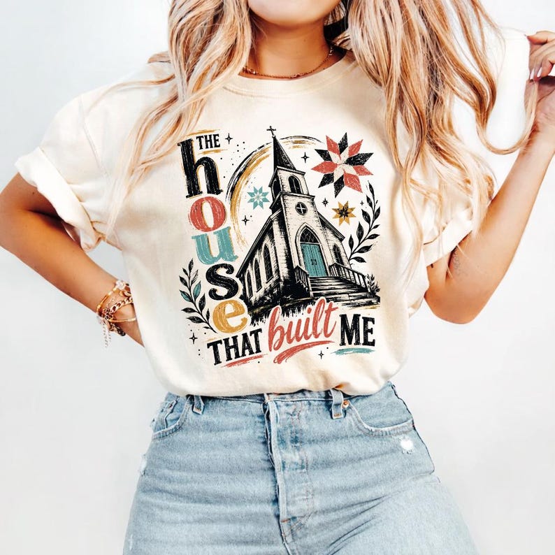 May include: A cream-colored short-sleeve t-shirt featuring a graphic of a church building with the text THE house THAT built ME. The design includes colorful star patterns, leafy branches, and decorative accents in shades of red, blue, and yellow.