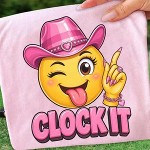 Puede incluir: Camiseta rosa con un emoji amarillo guiñando un ojo y con un sombrero de vaquero rosa. El emoji saca la lengua y hace un gesto con la mano con un anillo y una pulsera rosas. El texto "CLOCK IT" es rosa.