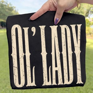May include: A black t-shirt featuring the text "OL' LADY" printed in a large, cream-colored, distressed font with a cracked texture. The lettering is vertically oriented and occupies the majority of the front of the garment.