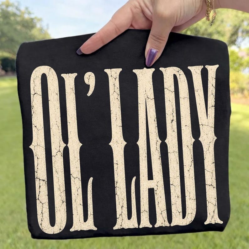 May include: A black t-shirt featuring the text "OL' LADY" printed in a large, cream-colored, distressed font with a cracked texture. The lettering is vertically oriented and occupies the majority of the front of the garment.