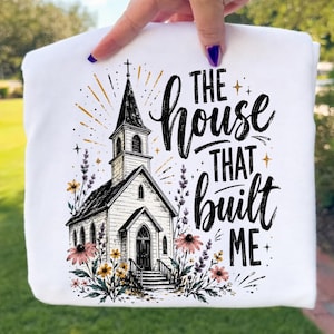 May include: A white t-shirt featuring a black ink illustration of a church building surrounded by colorful wildflowers. The text "THE HOUSE THAT BUILT ME" is printed in a bold, script-style font to the right of the church, accented by small gold stars.