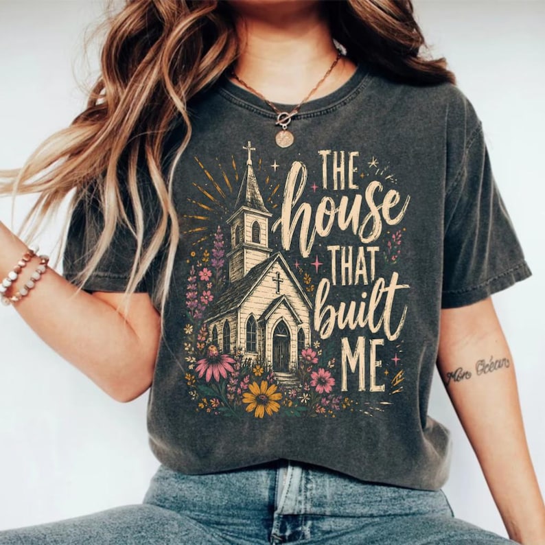 May include: A charcoal gray short-sleeve t-shirt featuring a graphic of a church building surrounded by colorful flowers. The text "THE house THAT built ME" is printed in a stylized script font next to the church illustration.