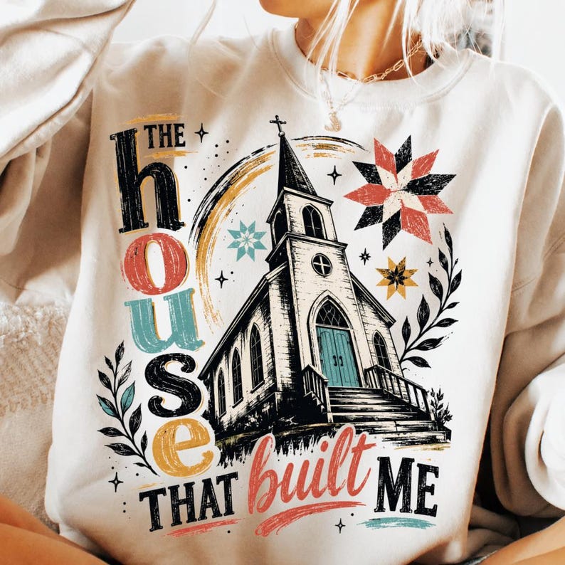 May include: A cream-colored sweatshirt featuring a graphic print of a church with a steeple and stairs. The text reads THE HOUSE THAT BUILT ME in black, red, and teal. Decorative elements include a star pattern, leaves, and a rainbow arc.
