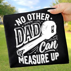 May include: A black t-shirt featuring a white graphic print of a tape measure and the text NO OTHER DAD CAN MEASURE UP. The design is centred on the fabric, utilising bold, stylised typography and a detailed illustration of a retractable tape measure.