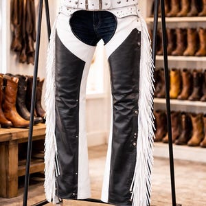 Puede incluir: Chaps de cuero en blanco y negro con flecos, colgados de un soporte de metal negro. Los chaps tienen un cinturón blanco con tachuelas plateadas y una hebilla. El diseño incluye un panel frontal blanco y paneles de pierna negros.