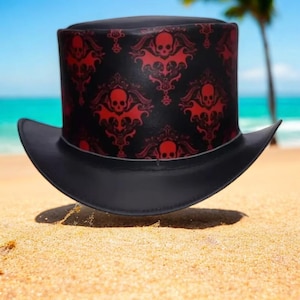 Op de afbeelding: Zwarte hoge hoed met een rood schedel- en vleermuismotief. De hoed heeft een brede rand en staat tegen een strandachtergrond met zand, zee en een palmboom. Het ontwerp van de hoed heeft een herhalend patroon van schedels en vleermuizen.