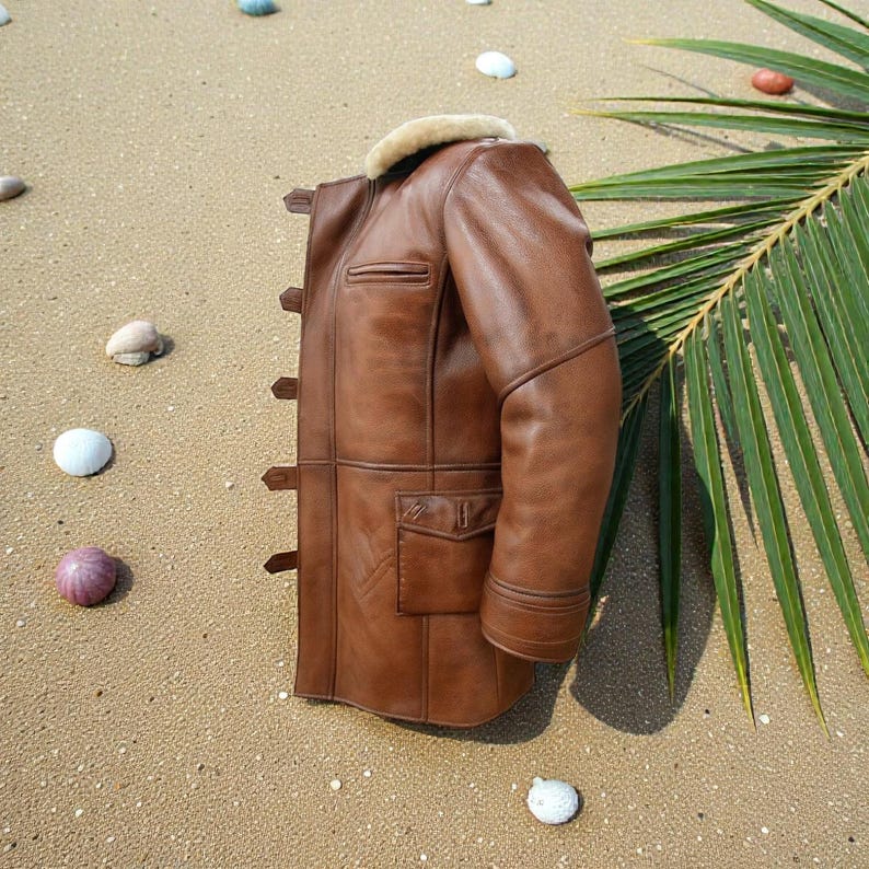 May include: A brown leather jacket featuring a cream-colored shearling collar, long sleeves, a vertical row of side toggle closures, and a rectangular flap pocket on the lower front section. The jacket has visible seam detailing throughout.