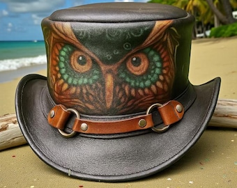 Puede incluir: Un sombrero de copa de cuero marrón oscuro con un rostro de búho pintado a mano en la parte frontal. El búho tiene ojos naranjas detallados rodeados de plumas verdes. Una correa de cuero color canela con dos anillos metálicos y remaches plateados rodea la base de la copa.