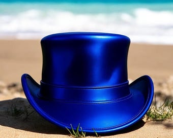 May include: A shiny, cobalt blue top hat with a wide brim sits on a sandy beach. The hat has a classic tall crown and a band around the base. The background features the ocean and a blue sky.