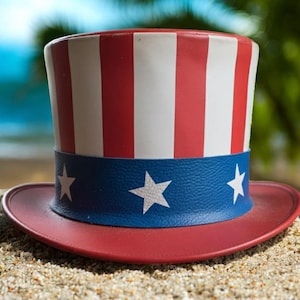 May include: A patriotic Uncle Sam hat in red, white, and blue. The hat has a wide brim and a crown with red and white stripes. A blue band with white stars encircles the base. The hat is on sand.
