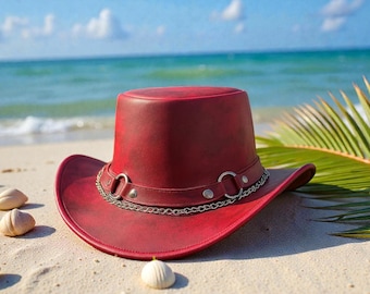 May include: A burgundy leather cowboy hat with a silver chain and buckle detail. The hat is on a sandy beach with seashells, palm leaves, and the ocean in the background. A stylish accessory.