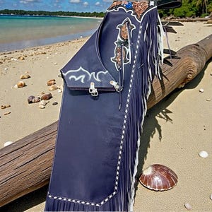 May include: Dark blue leather chaps with white fringe and decorative stitching. The chaps have detailed floral and scroll designs in brown and white. The chaps are on a log on a sandy beach with a seashell.