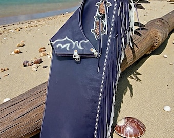 May include: Dark blue leather chaps with white fringe and decorative stitching. The chaps have detailed floral and scroll designs in brown and white. The chaps are on a log on a sandy beach with a seashell.