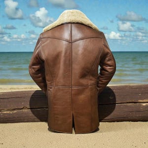 May include: A brown leather jacket featuring a thick cream-colored shearling collar. The jacket has a paneled construction with visible seams, long sleeves, and a straight hemline with a center back vent, presented from a rear view.