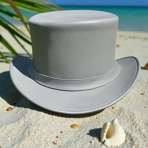 Puede incluir: Un sombrero de copa gris con una corona redondeada y un ala ancha. El sombrero está hecho de un material liso y tiene una banda alrededor de la base de la corona. El sombrero está en una playa de arena con una concha en primer plano.