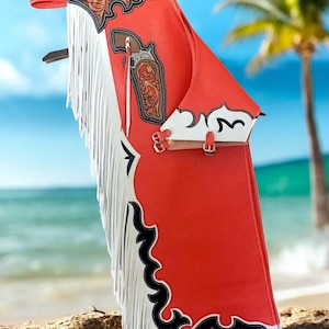 May include: Red leather chaps with white fringe and decorative tooling. The chaps feature a brown leather holster design and black and white decorative trim. The chaps are designed with buckles and straps.