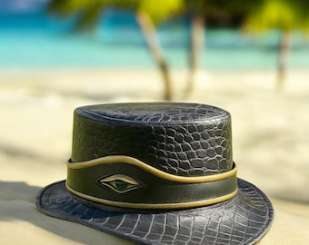Peut inclure: Un chapeau en cuir noir avec un motif texturé en forme de crocodile. Le chapeau présente une bande noire avec une bordure dorée et un motif en forme d'œil vert. Le chapeau est posé sur une surface sablonneuse avec une plage floue et des palmiers en arrière-plan.