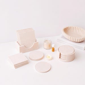 May include: A collection of light beige home decor items including a set of square coasters in a holder, a set of round coasters in a holder, a small glass jar with a lid, a tiny amber glass bottle, a tea light candle, and a ribbed oval bowl.