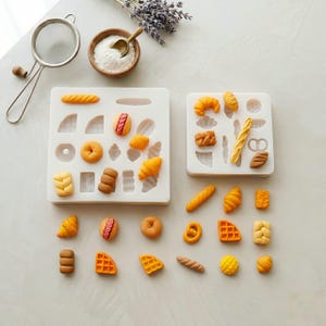 May include: Miniature baked goods, such as croissants and bagels, are arranged on a white surface. The scene includes silicone moulds and a small bowl of white powder, suggesting a baking theme. The colors are warm and inviting, with a focus on the food.