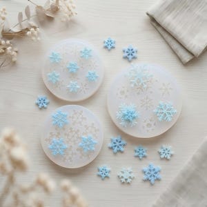 May include: Three circular translucent silicone molds featuring various snowflake patterns, accompanied by several small, light blue, three-dimensional snowflake-shaped resin or clay pieces scattered around the molds on a light surface.
